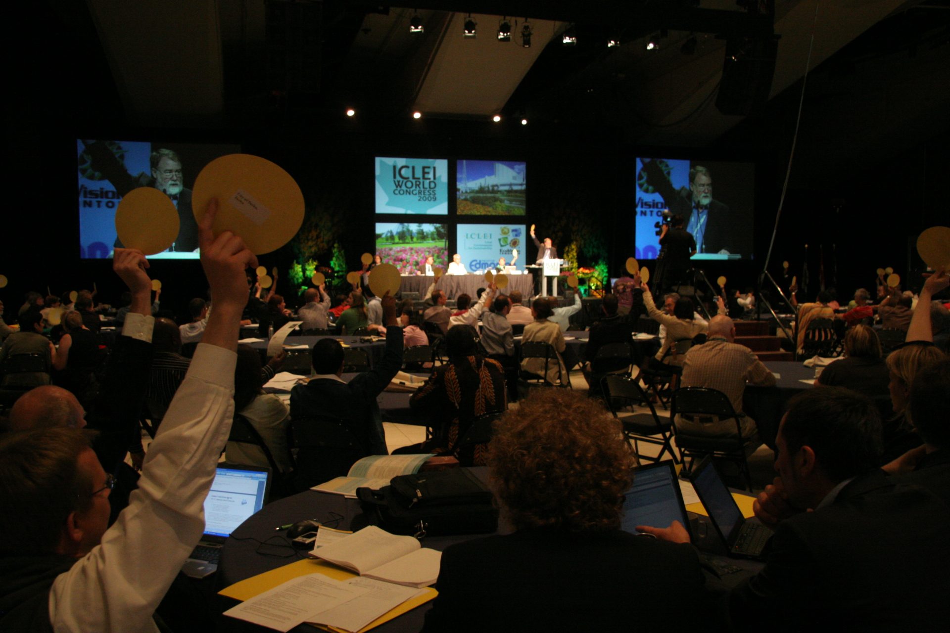 A shot of a the audience at the ICLEI World Congress, some with hands in the air holding a round disc, all facing the professional panelists on the stage.